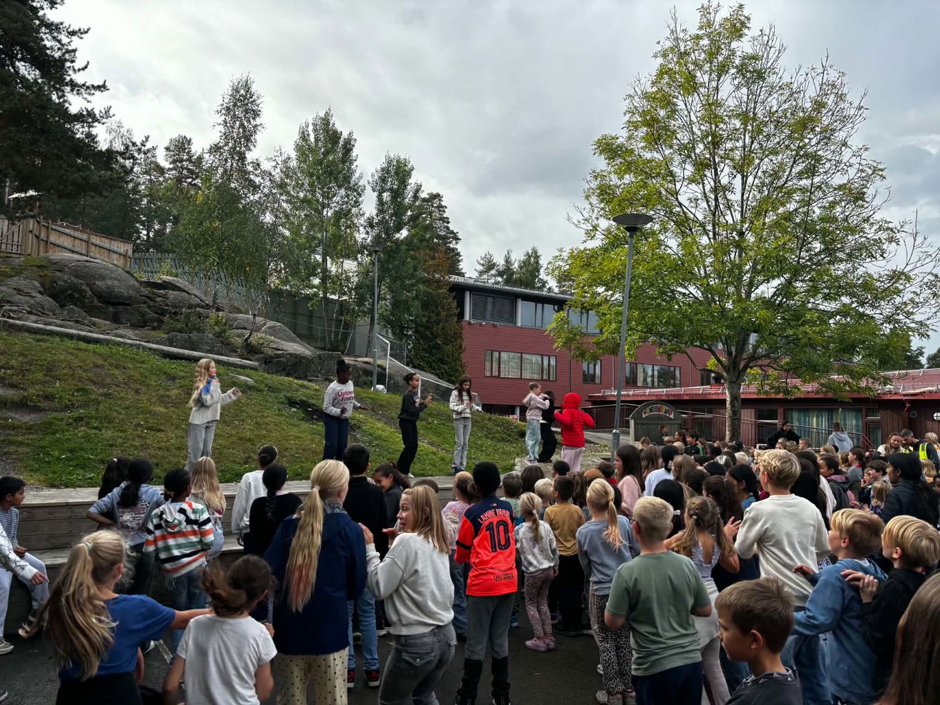 en gruppe mennesker som står på en gresskledd ås med trær og bygninger i bakgrunnen