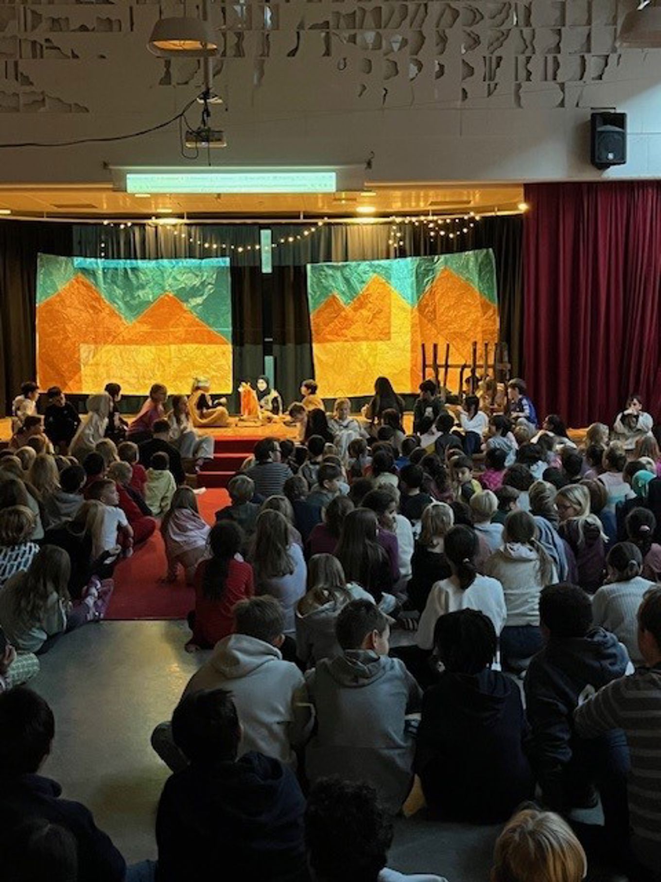 en gruppe mennesker som sitter i et rom med en scene og en stor skjerm en gruppe mennesker som sitter i et rom med en scene og en stor skjerm