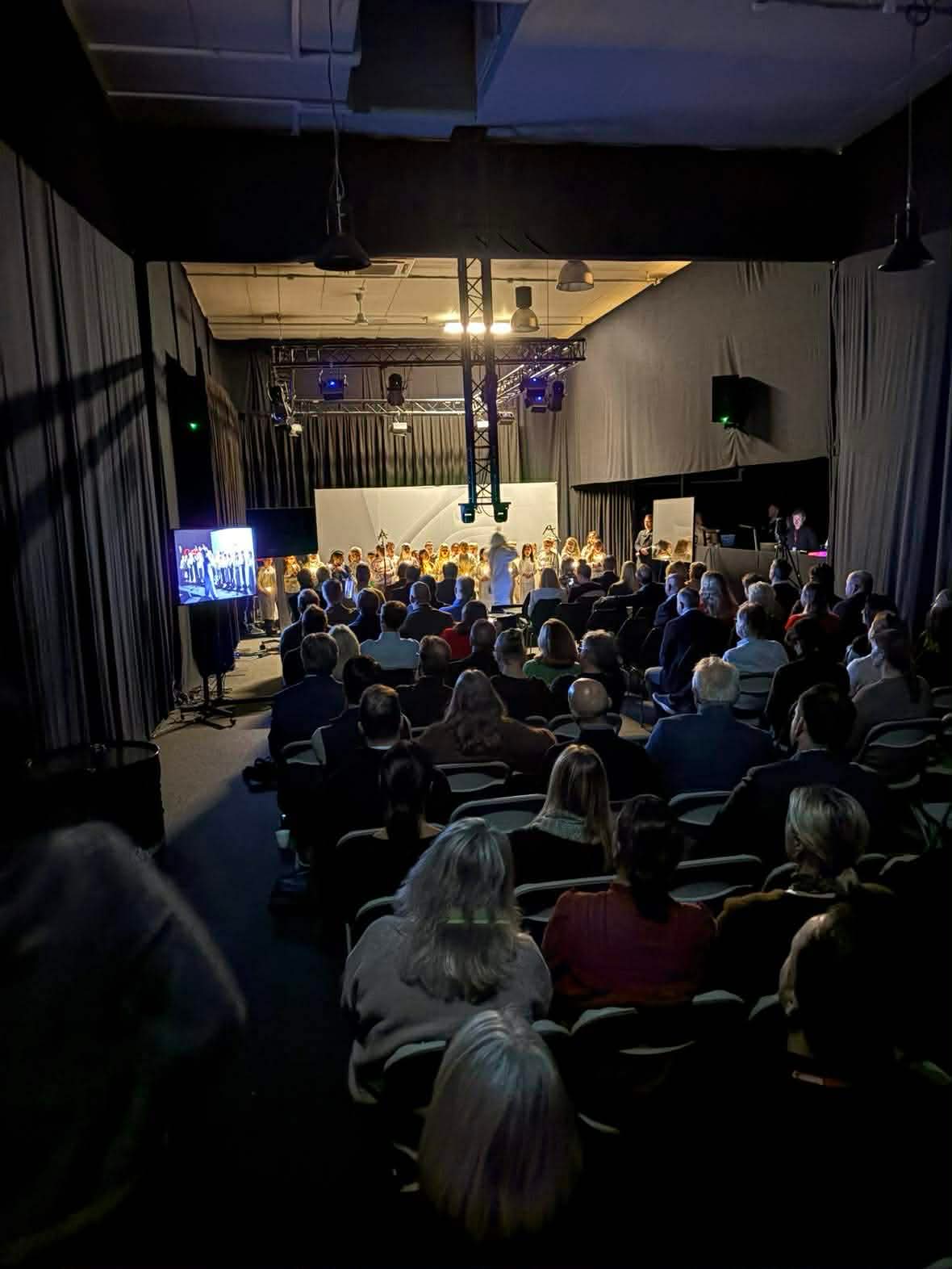 en gruppe mennesker som sitter i et rom med en scene og en stor skjerm